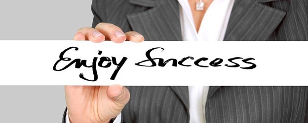 Says "Enjoy Success" in black letters with white background with a business person in professional dress behind the sign